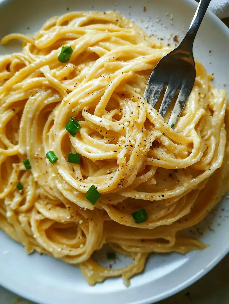 Cerca de pasta cremosa con cebolla verde y pimienta en plato blanco, con tenedor dentro de los fideos.
