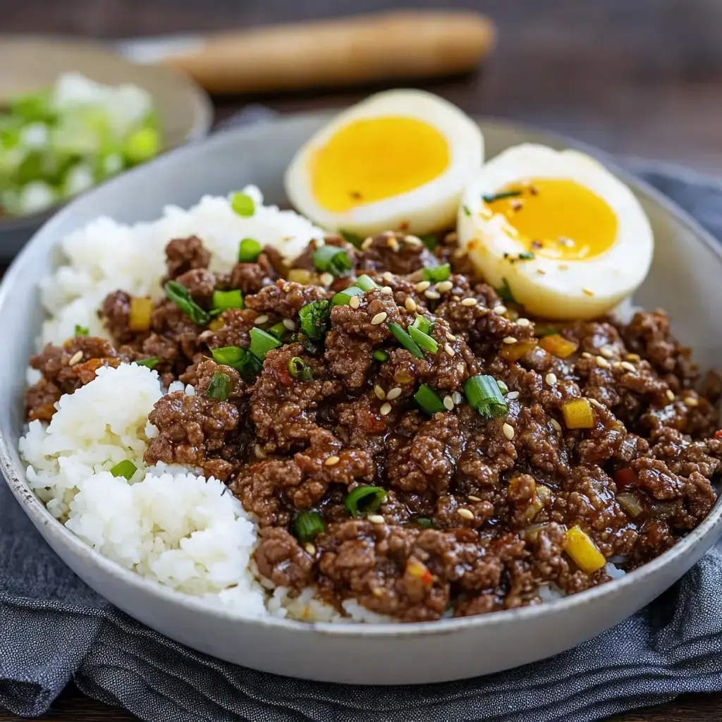 Un tazón de arroz cubierto con carne molida sabrosa, decorado con cebollines y semillas de sésamo, junto a dos mitades de huevo suave.