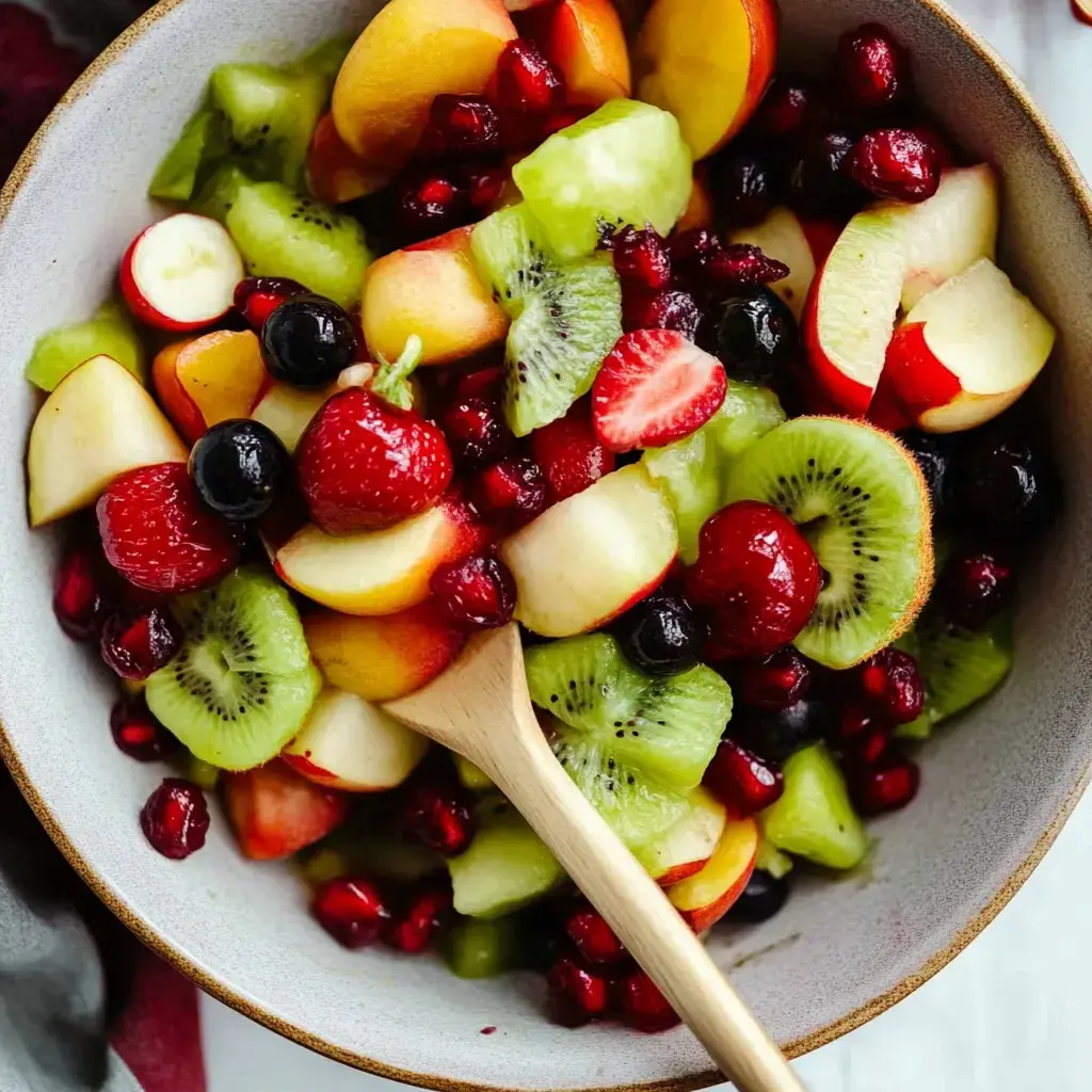Una vibrante ensalada de frutas que incluye rodajas de manzanas, fresas, kiwi y melones, mezclados con frambuesas y granada en un bol con cuchara de madera.