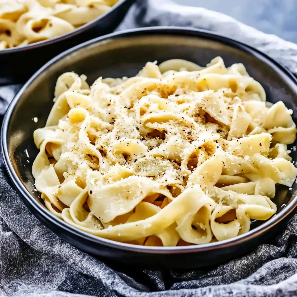 Un plato de pasta tipo fettuccine cubierto con queso rallado y pimienta, servido sobre un mantel gris texturizado.
