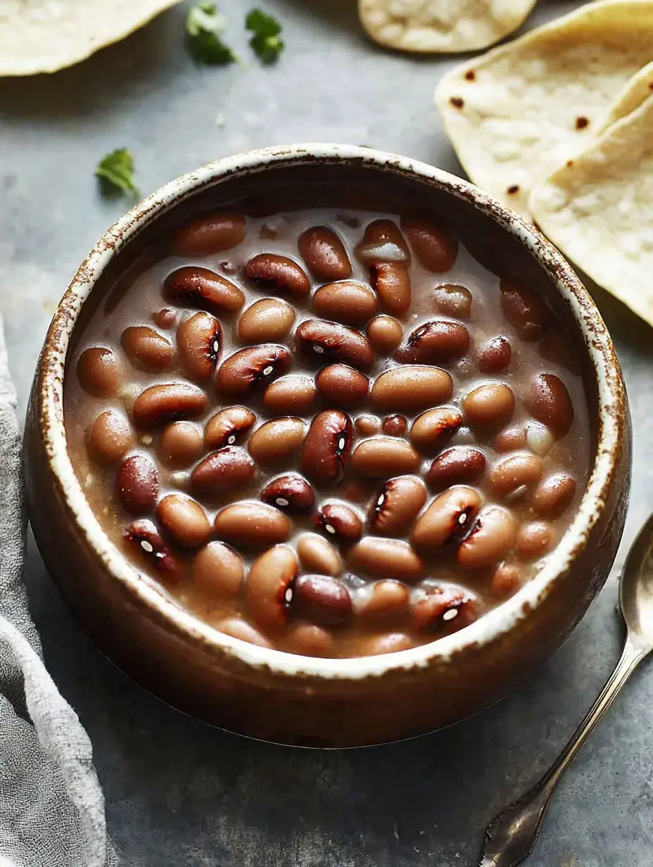 Un plato de frijoles en caldo rodeado de tortillas y cilantro fresco sobre una mesa de madera.