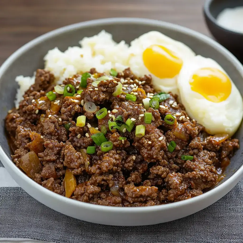 Un plato de carne molida sazonada con cebolleta y semillas de sésamo, acompañado de arroz y dos huevos de yema blanda.
