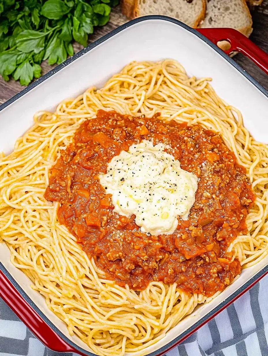Un primer plano de espagueti cubierto con salsa de carne y una buena cucharada de salsa cremosa, en una bandeja cuadrada, decorada con perejil fresco y un pan al fondo.