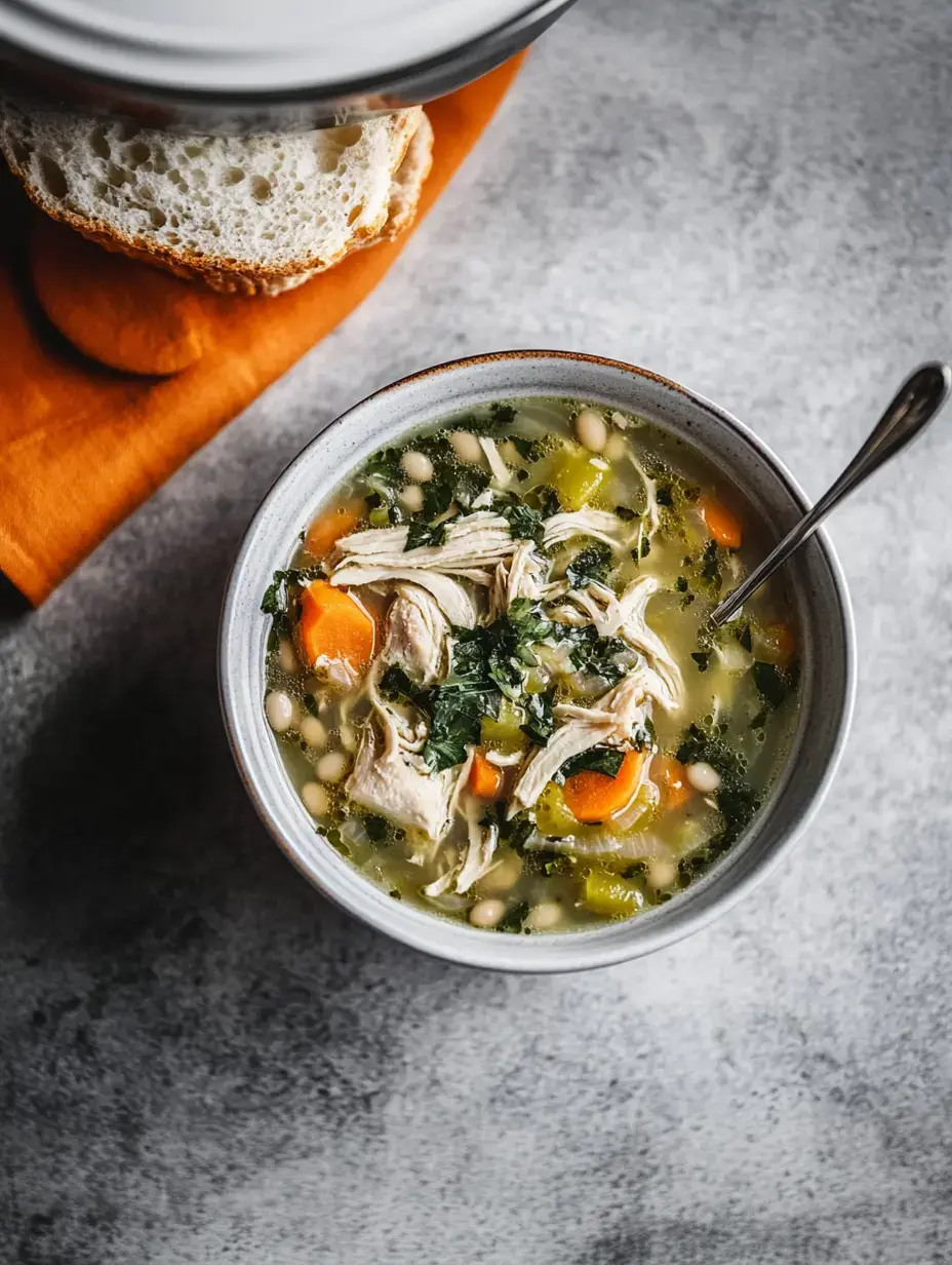 Un plato de sopa de pollo con pollo desmenuzado, zanahorias, verduras de hoja y alubias blancas, al lado de una rebanada de pan sobre una mesa gris.