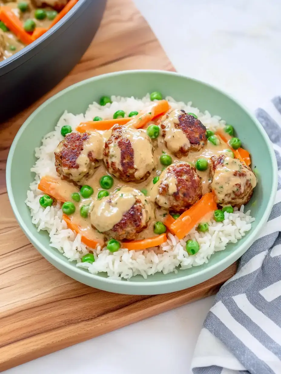 Un plato con arroz, albóndigas en salsa cremosa, guisantes y tiras de zanahoria, servido en un cuenco verde.
